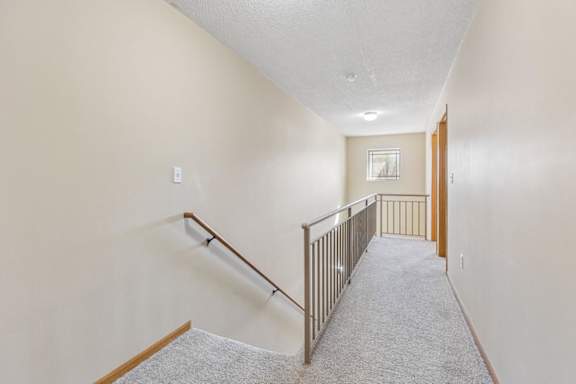 Upper-level apartment hallway with neutral carpet, stair railing, and natural light from a distant window.