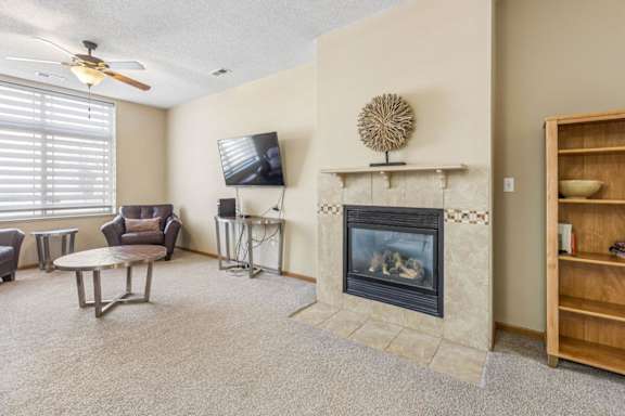 Cozy apartment living room with neutral carpet, tiled fireplace, large window with blinds, ceiling fan, and seating area with a wall-mounted TV.