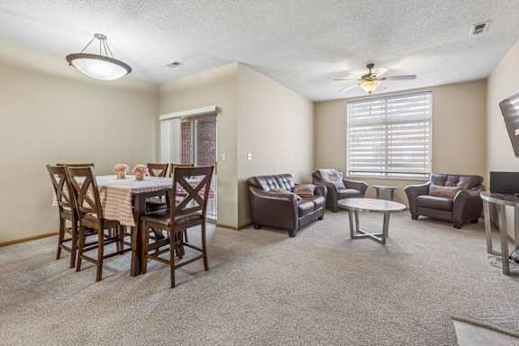 Spacious apartment living and dining area with neutral carpet, large window with blinds, ceiling fan, and sliding glass door leading to a patio.