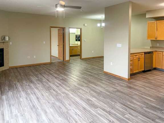 a living room with a corner fireplace, luxury vinyl tile flooring, and a ceiling fan next to a kitchen