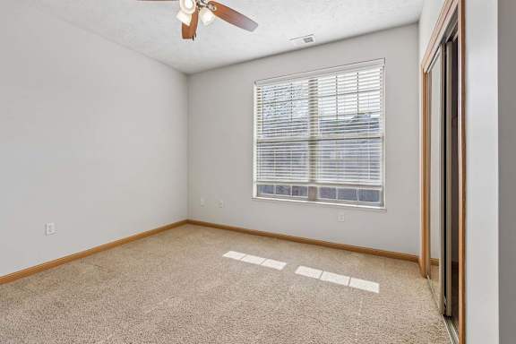 Sunlit apartment bedroom with a wide window, mirrored sliding closet doors, neutral carpeting, and a ceiling fan.