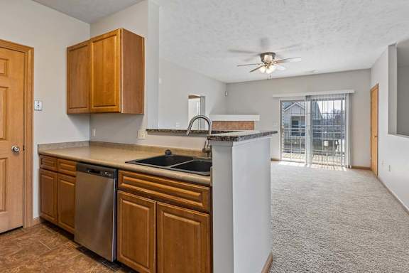 Open-concept apartment kitchen and living area with wood cabinetry, breakfast bar, dishwasher, ceiling fan, and sliding glass door.