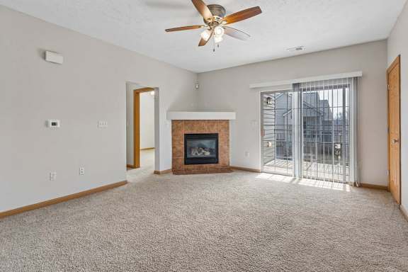 Bright, empty apartment living room with beige carpet, tiled fireplace, ceiling fan, and sliding glass doors opening to a balcony.