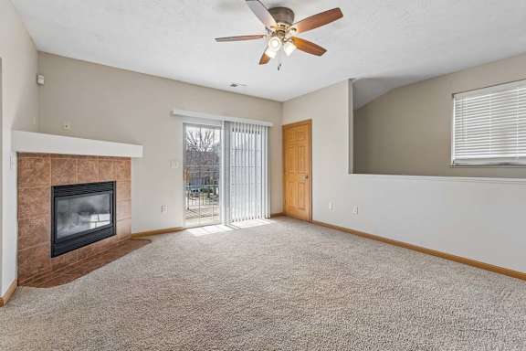 Bright apartment living room with beige carpet, tiled fireplace, ceiling fan, and sliding glass doors leading to a balcony.