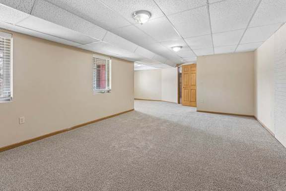 Spacious, empty lower-level room with neutral carpet, drop ceiling, recessed lighting, windows with blinds, and white brick wall.