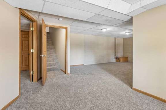Empty lower-level room with neutral carpet, drop ceiling, white brick walls, overhead lighting, and stair access.