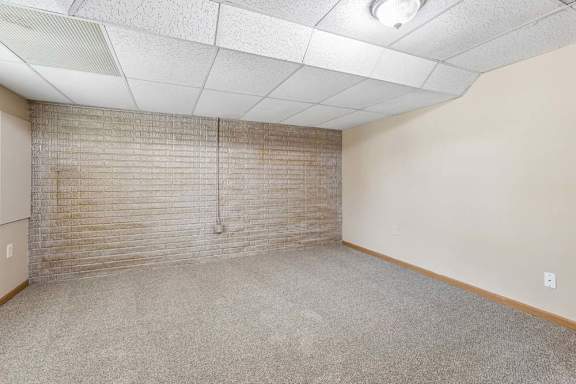 Empty lower-level room with neutral carpet, exposed brick accent wall, drop ceiling, and overhead lighting.