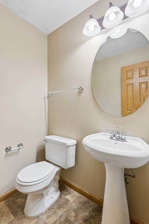 Half bathroom with pedestal sink, oval mirror, neutral tile flooring, bright vanity lighting, and towel bars.