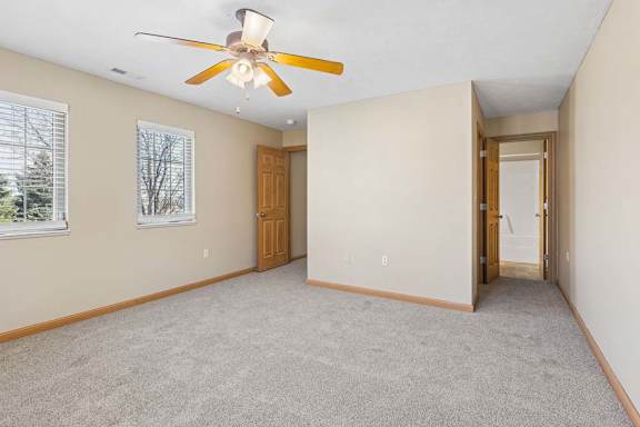 Bright, empty bedroom with neutral carpet, two windows with blinds, ceiling fan, wood trim, and direct bathroom access.