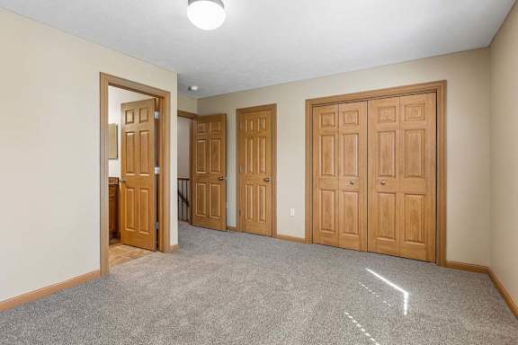 Empty bedroom with neutral carpet, double closet doors, wood trim, overhead lighting, and direct bathroom access.