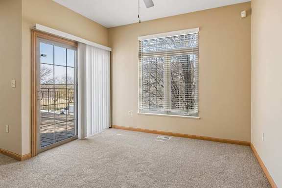 Bright, empty living area with neutral carpet, large window with blinds, ceiling fan, and sliding glass door to the deck.