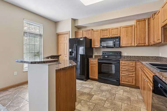 Kitchen with wood cabinetry, speckled countertops, black appliances, breakfast bar, tile flooring, and a large window.