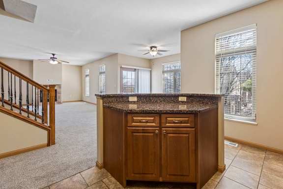 Open kitchen and living area with wood cabinetry, speckled breakfast bar, tile and carpet flooring, large windows, and staircase.