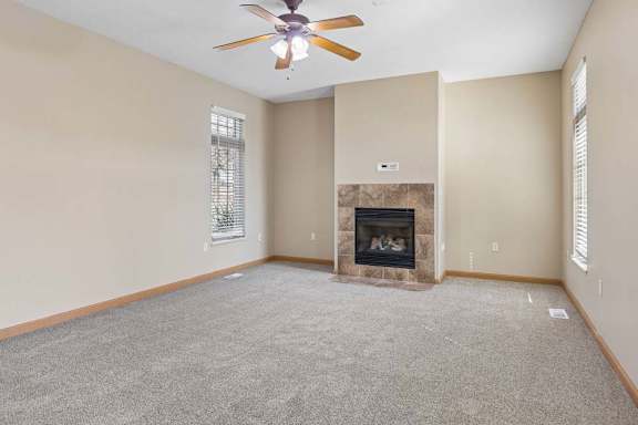 Bright, empty living room with neutral carpet, tiled fireplace, ceiling fan, and tall windows with blinds.