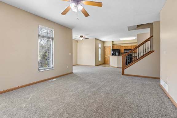 Spacious, empty living area with neutral carpet, ceiling fans, tall windows, open kitchen, and wood-railed staircase.