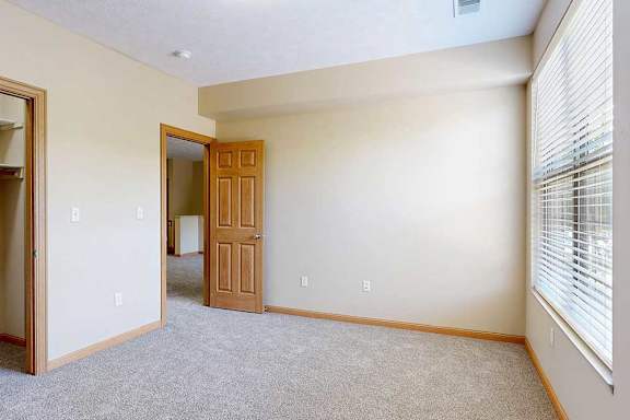 A carpeted bedroom with a large window from plenty of natural light, and an open door to the living room.