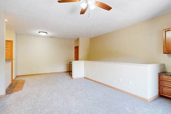 A carpeted living room with a ceiling fan and a half wall separating the stairs down to the door.
