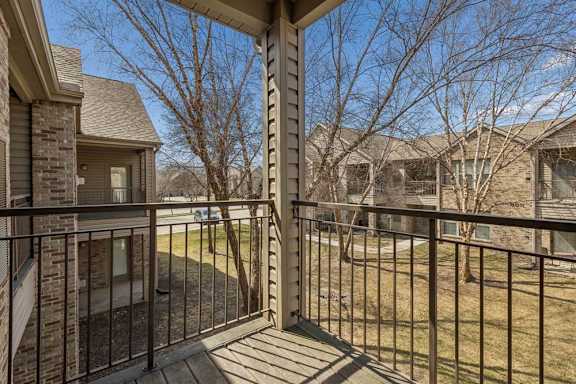 Covered apartment balcony with metal railing overlooking mature trees, landscaped grounds, and neighboring community buildings.