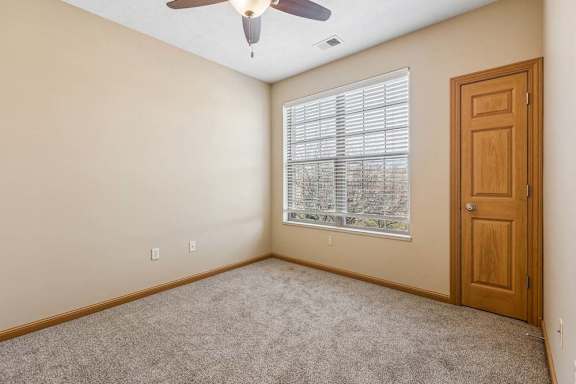 Bright apartment bedroom with a wide window and blinds, neutral carpeting, ceiling fan, and a wood closet door.