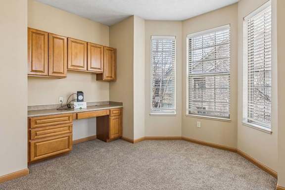 Bright apartment office nook with built-in wood cabinetry, a desk area, carpeted flooring, and three large windows with blinds.