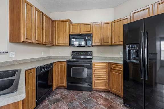 U-shaped apartment kitchen with extensive wood cabinetry, wraparound counters, black appliances, and a double-basin sink.