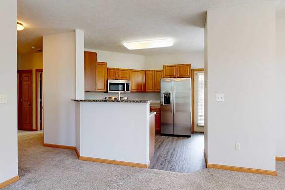 A kitchen with a breakfast bar countertop, wooden cabinets, and stainless steel appliances next to a living room.