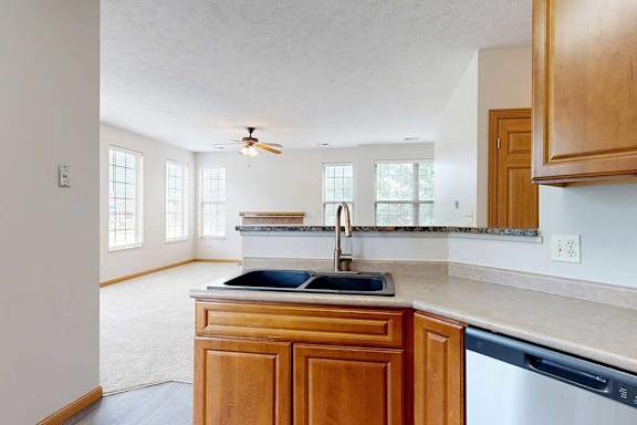 A kitchen with wooden cabinets, and a double basin stone sink, with the living room in the background.