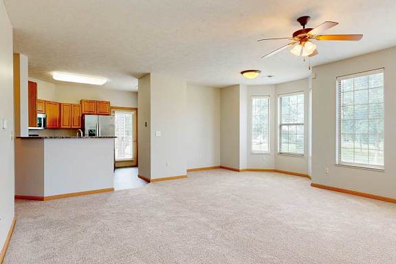 A spacious living room with a carpeted floor, a bay window in the dining area, a ceiling fan, and a breakfast bar countertop to the kitchen.