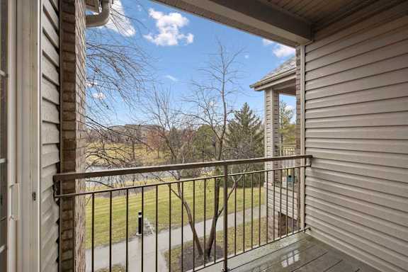 Covered balcony with metal railing and views of green space and neighboring buildings.