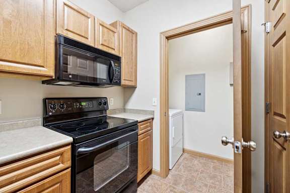 Kitchen with black refrigerator, oven, and microwave, natural wood cabinets, and tile flooring.