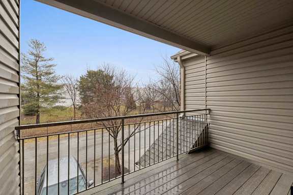Covered apartment balcony with wood-look decking and metal railing, overlooking a parking area and mature trees.