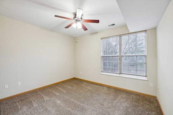 Bright bedroom with plush carpet, neutral walls, ceiling fan with light, and large window with blinds.