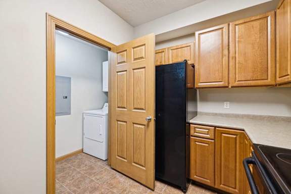 Apartment kitchen with wood cabinetry and laminate countertops, featuring a black refrigerator and adjacent in-unit laundry closet with washer and dryer.