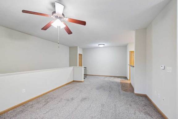 Spacious living area with plush carpet, neutral walls, ceiling fan, and half wall overlooking stairs