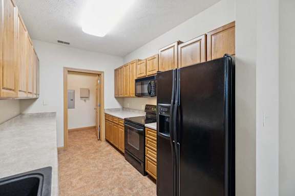Galley-style kitchen with natural wood cabinets, black refrigerator, oven and microwave, laminate countertops, and tile flooring.