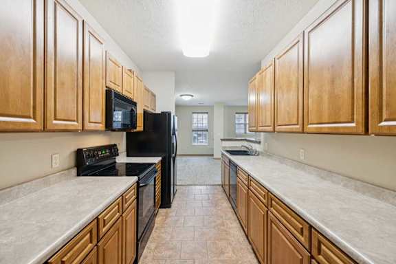 Galley-style kitchen with natural wood cabinets, laminate countertops, black oven and microwave, and tile flooring leading to open living area.