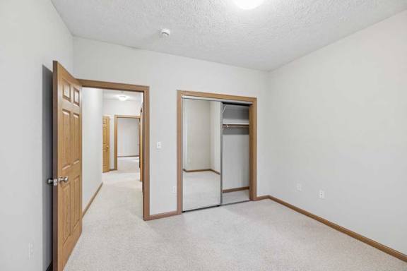 Bedroom with mirrored closet doors, neutral carpet, and hallway access in a well-lit apartment space.