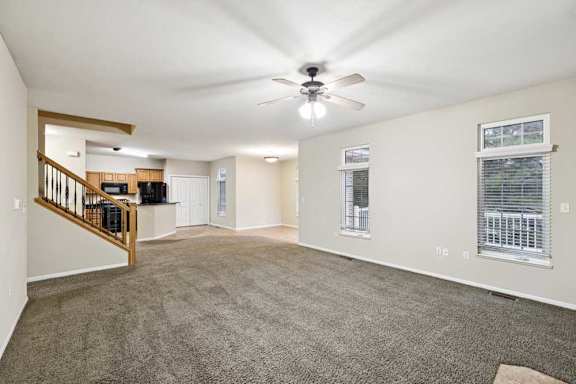 Spacious open living area with carpet flooring, staircase with wood railing, and kitchen in background.