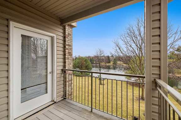 Private apartment balcony with wood decking and metal railing overlooking a pond and landscaped green space, accessed by a glass patio door.