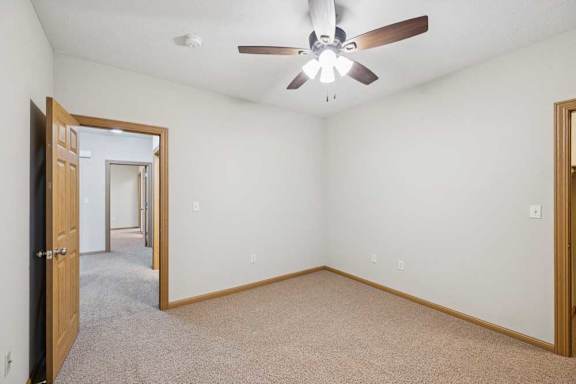 Empty apartment bedroom with neutral carpet, white walls, wood trim, and ceiling fan, connected to an adjoining hallway.