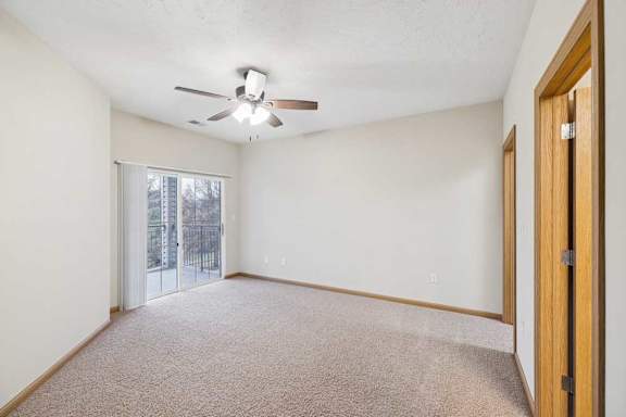 Spacious apartment living room with neutral carpet, ceiling fan, and sliding glass door leading to a private balcony with outdoor views.