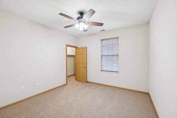 Empty bedroom with neutral carpet, ceiling fan, wood trim, and open doorway to an attached closet.