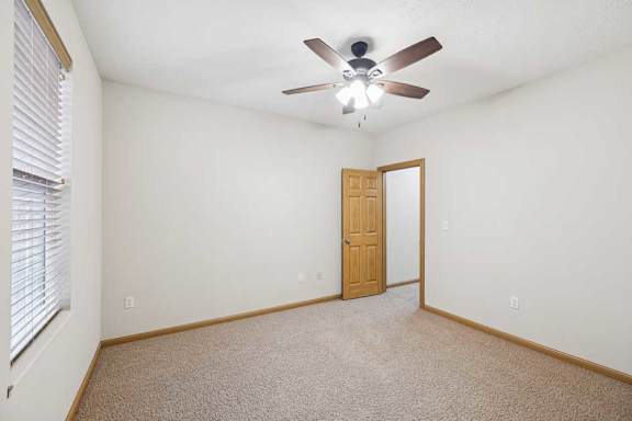 Bright, empty apartment bedroom with neutral carpet, white walls, wood trim, a ceiling fan with light, and a large window with blinds for natural light.