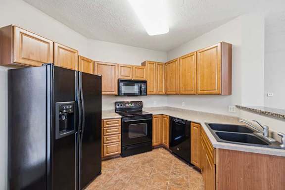 U-shaped apartment kitchen with wood cabinetry, black refrigerator, oven and microwave, laminate countertops, and tile flooring.
