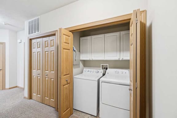 In-home laundry closet with side-by-side washer and dryer, upper storage cabinets, tile flooring, and wood bi-fold doors in a carpeted hallway.