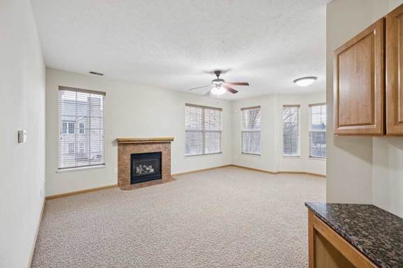 Open living room interior with neutral carpet, multiple windows, ceiling fan, and a corner fireplace with tile surround and wood mantel.