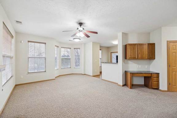 Open apartment living and dining area with neutral carpet, multiple windows, ceiling fan, and built-in desk with upper cabinets near the kitchen.