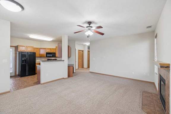 Open-concept living room with neutral carpet, ceiling fan, corner fireplace with tile surround, and view into the adjoining kitchen.