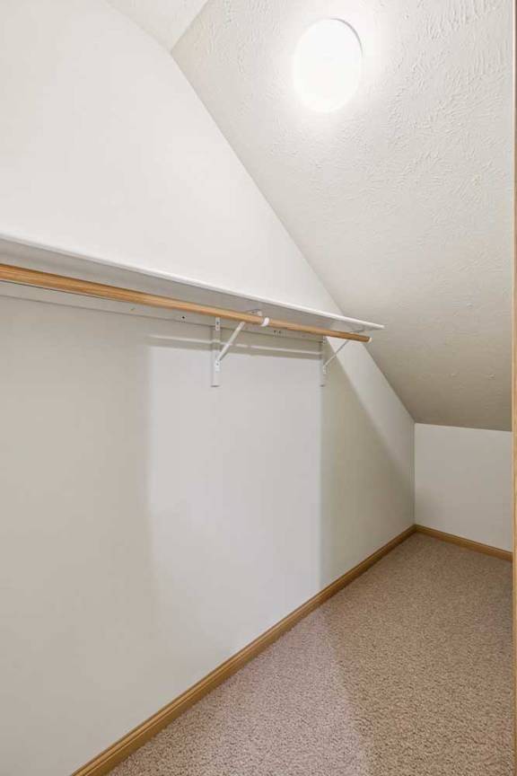 Under-stair storage space with sloped ceiling, carpet flooring, hanging rod, and built-in shelf for organized storage.