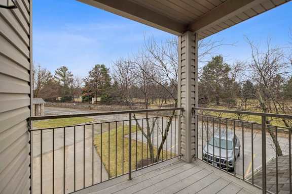 Private apartment balcony with wood decking and metal railing overlooking a parking area and landscaped grounds with mature trees.